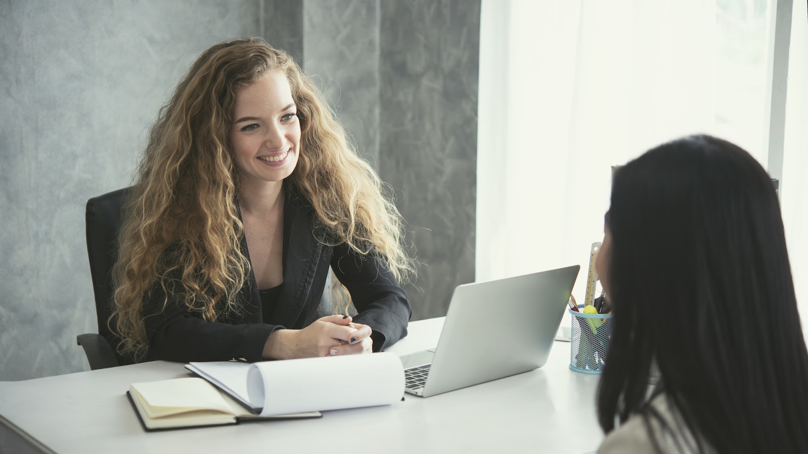 Woman sitting at desk smiling and talking to another woman